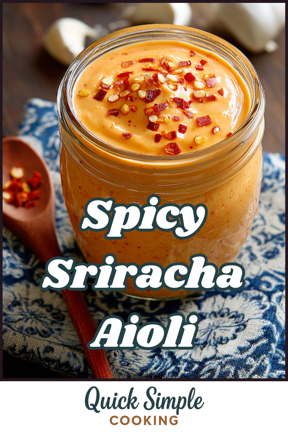 A jar of Spicy Sriracha Aioli topped with red pepper flakes sits on a blue-patterned cloth, with a wooden spoon holding more flakes beside it.