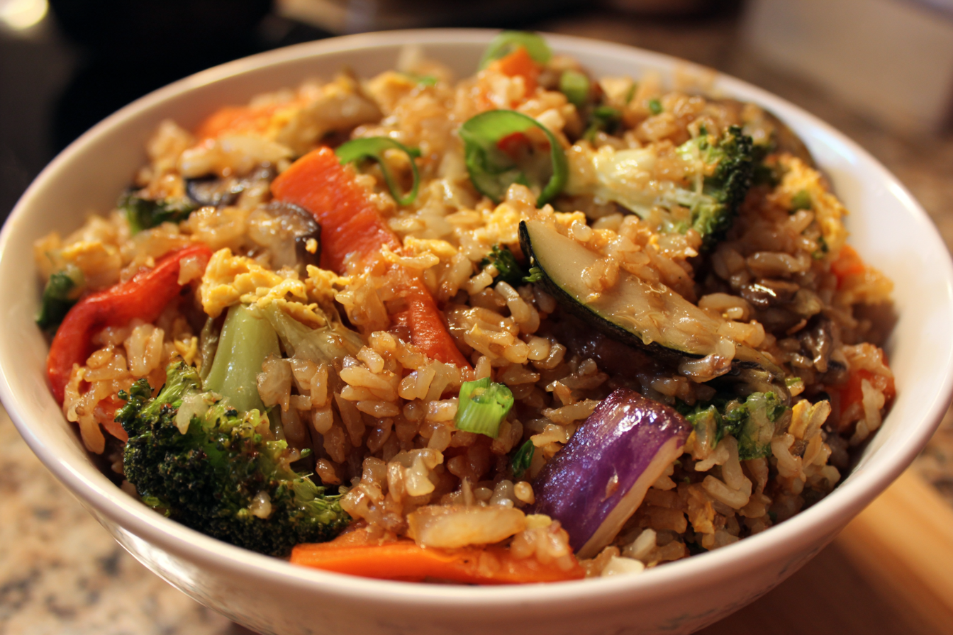 A bowl of fried rice with assorted vegetables including broccoli, red bell pepper, carrots, mushrooms, and egg, garnished with sliced green onions.