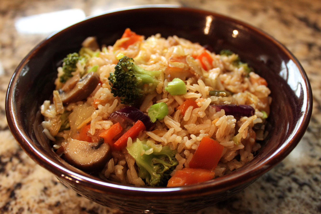 A brown bowl filled with vegetable fried rice, including broccoli, mushrooms, carrots, and onions, on a granite countertop.